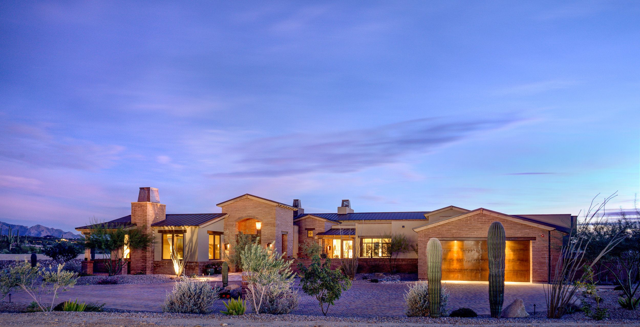 Long low elevation of the ranch house at twilight, warm interior light spilling across desert landscaping