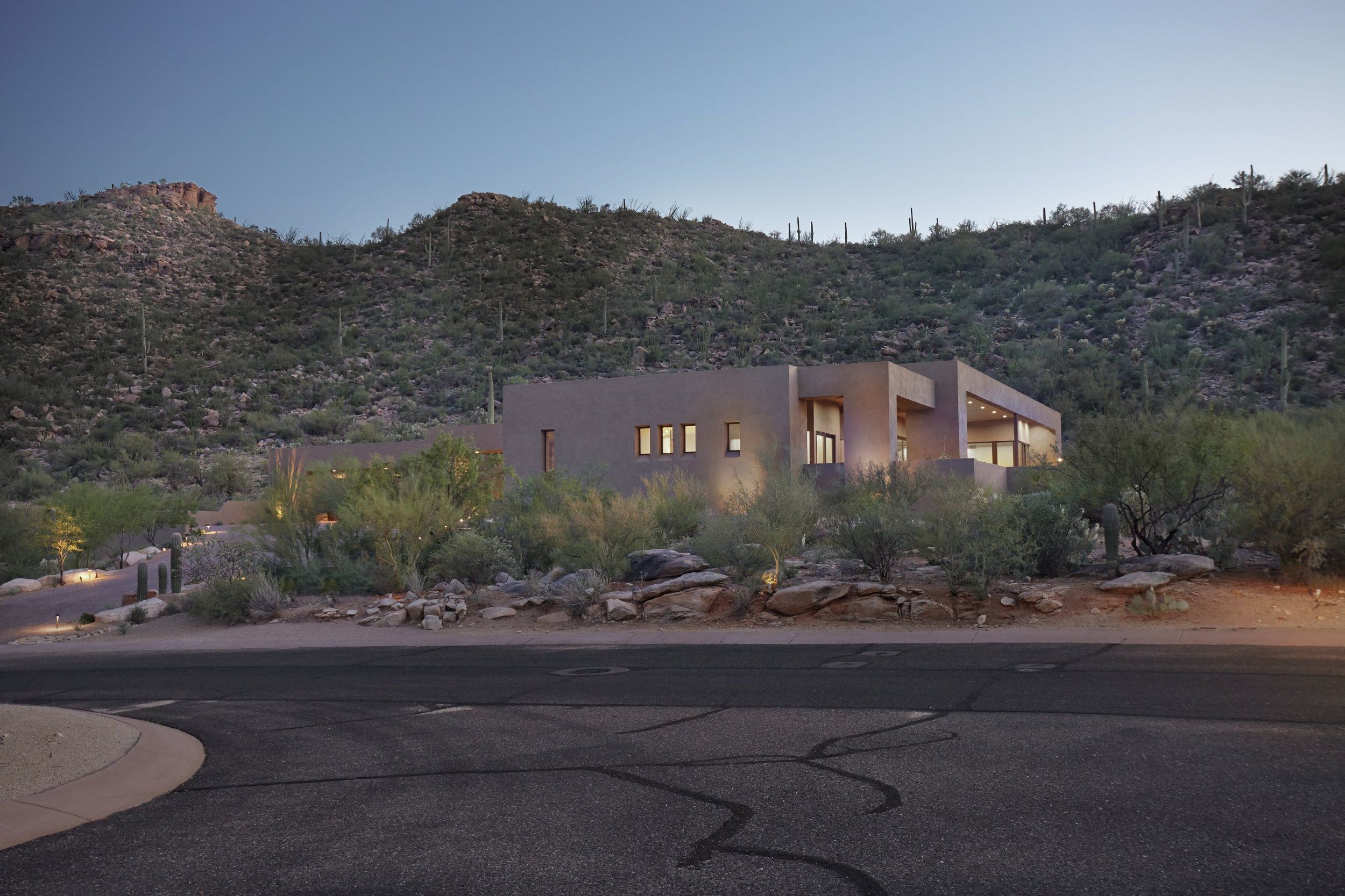 Street view of the low stucco home set against a desert hillside at dusk